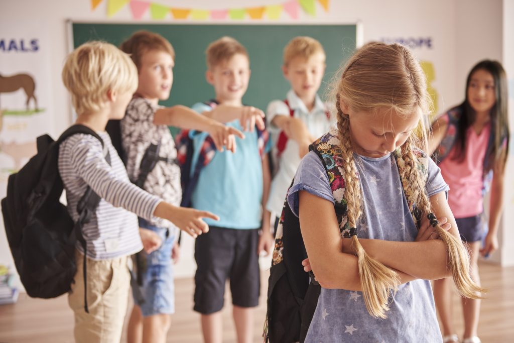 A young girl in a classroom looks dejected as her classmates point and laugh at her.