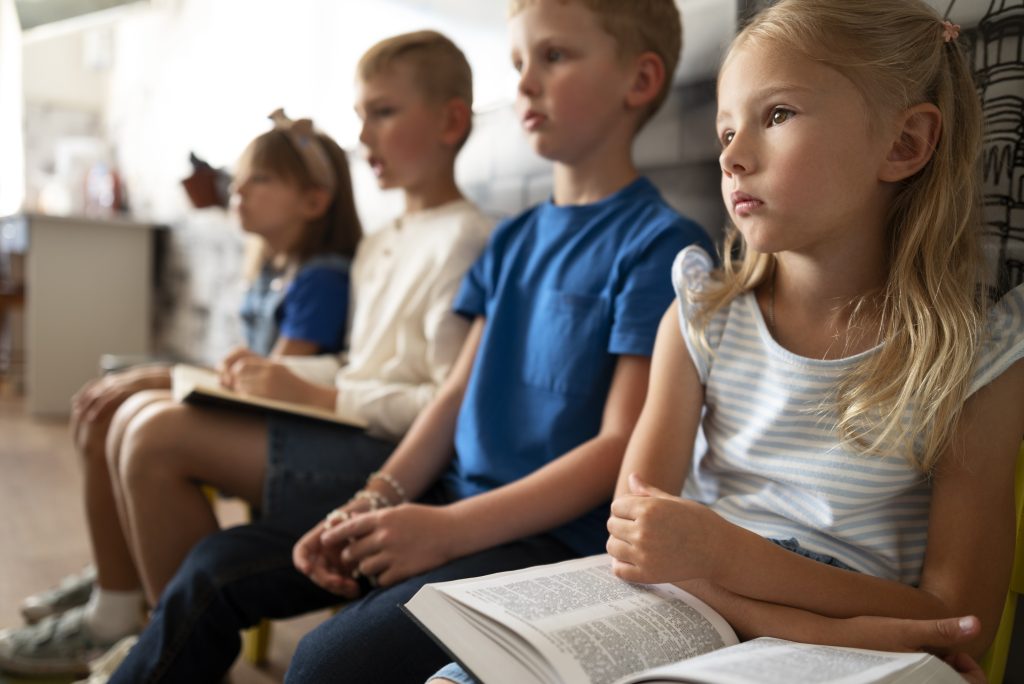 A row of children sit quietly with textbooks and hands in laps.