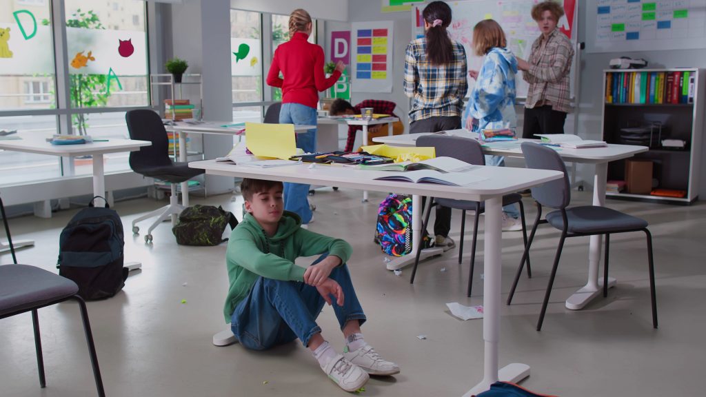 A boy hides under a desk in a classroom, looking like an overstimulated neurodivergent learner .