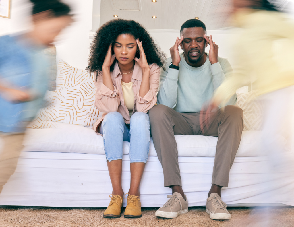 Parents of PDA in ADHD and autistic kids sit on couch looking frustrated while kids run around them